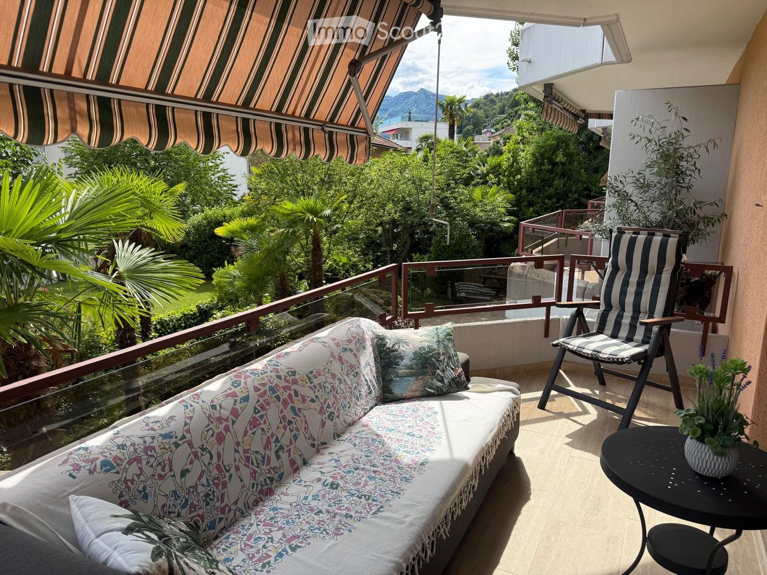 Balcony with a sofa, chair, small table, and potted plant, overlooking a lush, green landscape with palm trees and mountains.