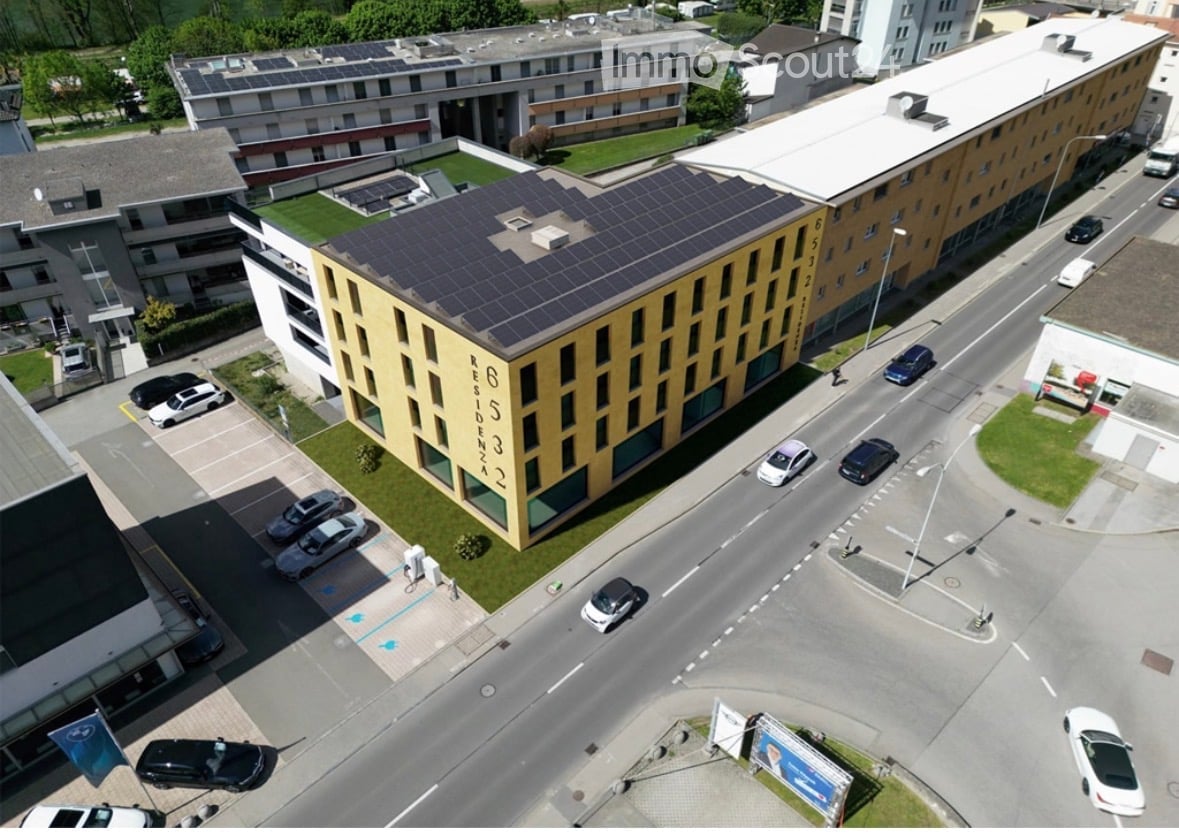 Aerial view of a modern multi-story residential building with solar panels on the roof, surrounded by parked cars and greenery.