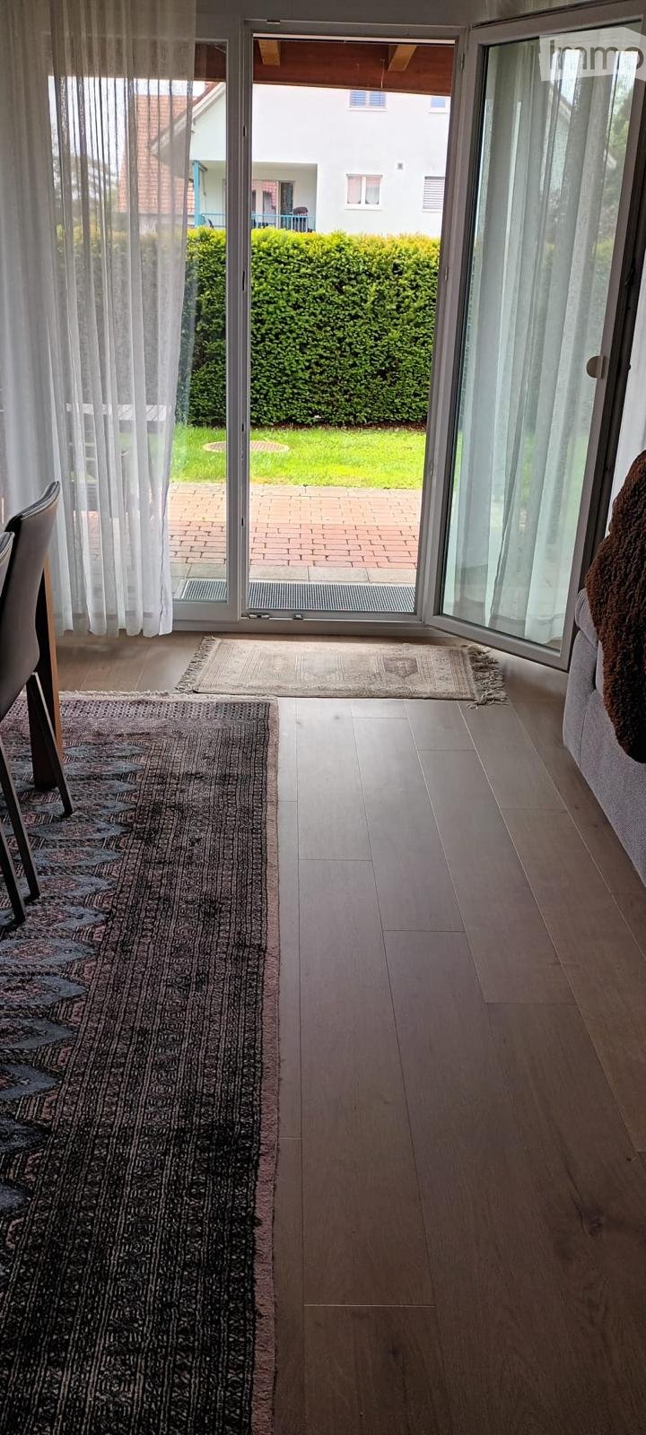 Bright hallway with wooden flooring, large sliding glass doors leading to a garden, patterned rugs, and a visible chair and sofa.