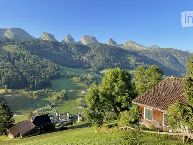 Scenic mountain landscape with a small village nestled in a lush green valley, surrounded by dense forests and towering peaks under a clear blue sky.