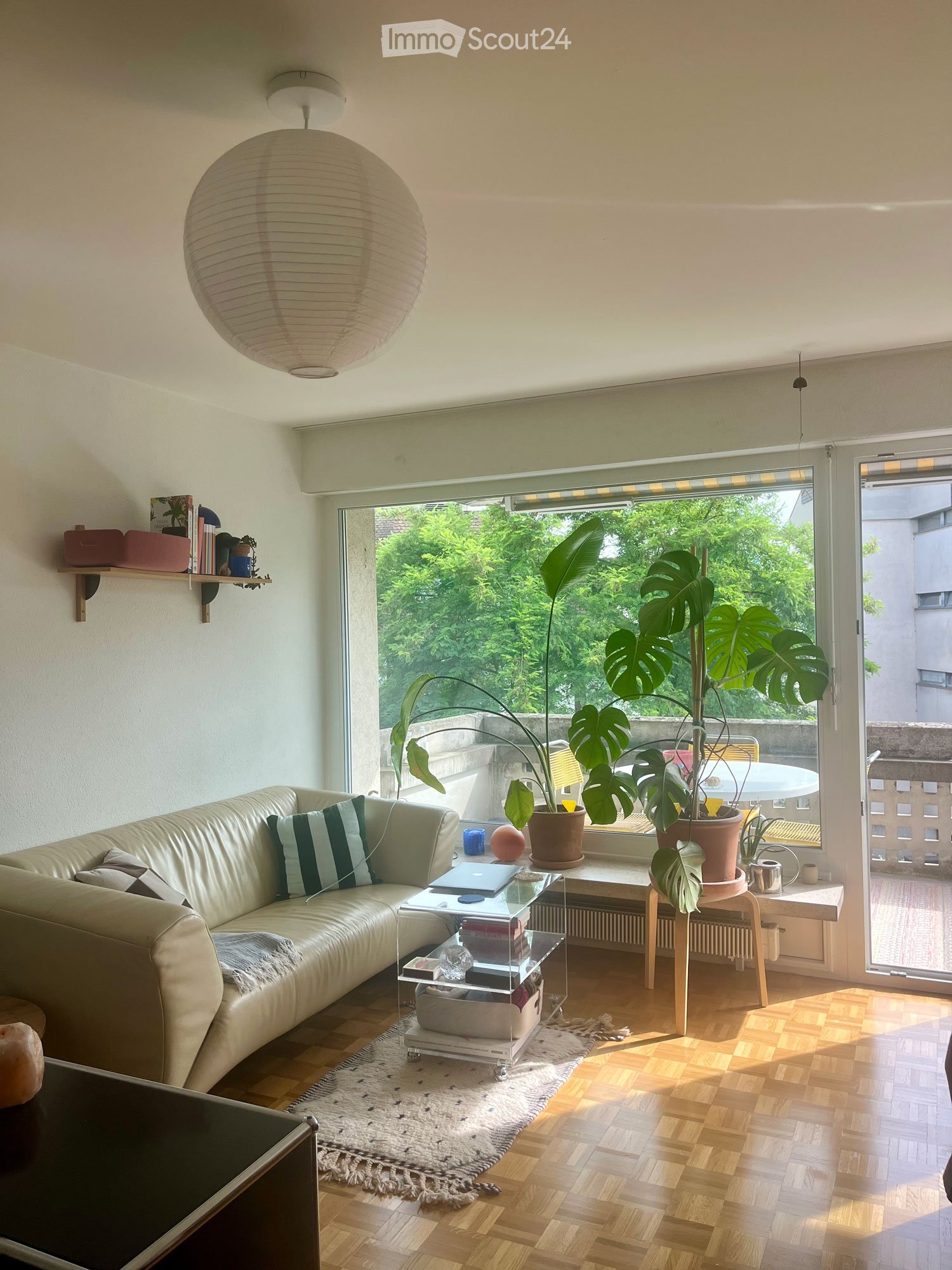 Bright living room with wooden flooring, large window offering a view of greenery, a cream-colored sofa with striped cushions, a glass coffee table with a laptop, a potted plant, and a shelf with books and decorative items.