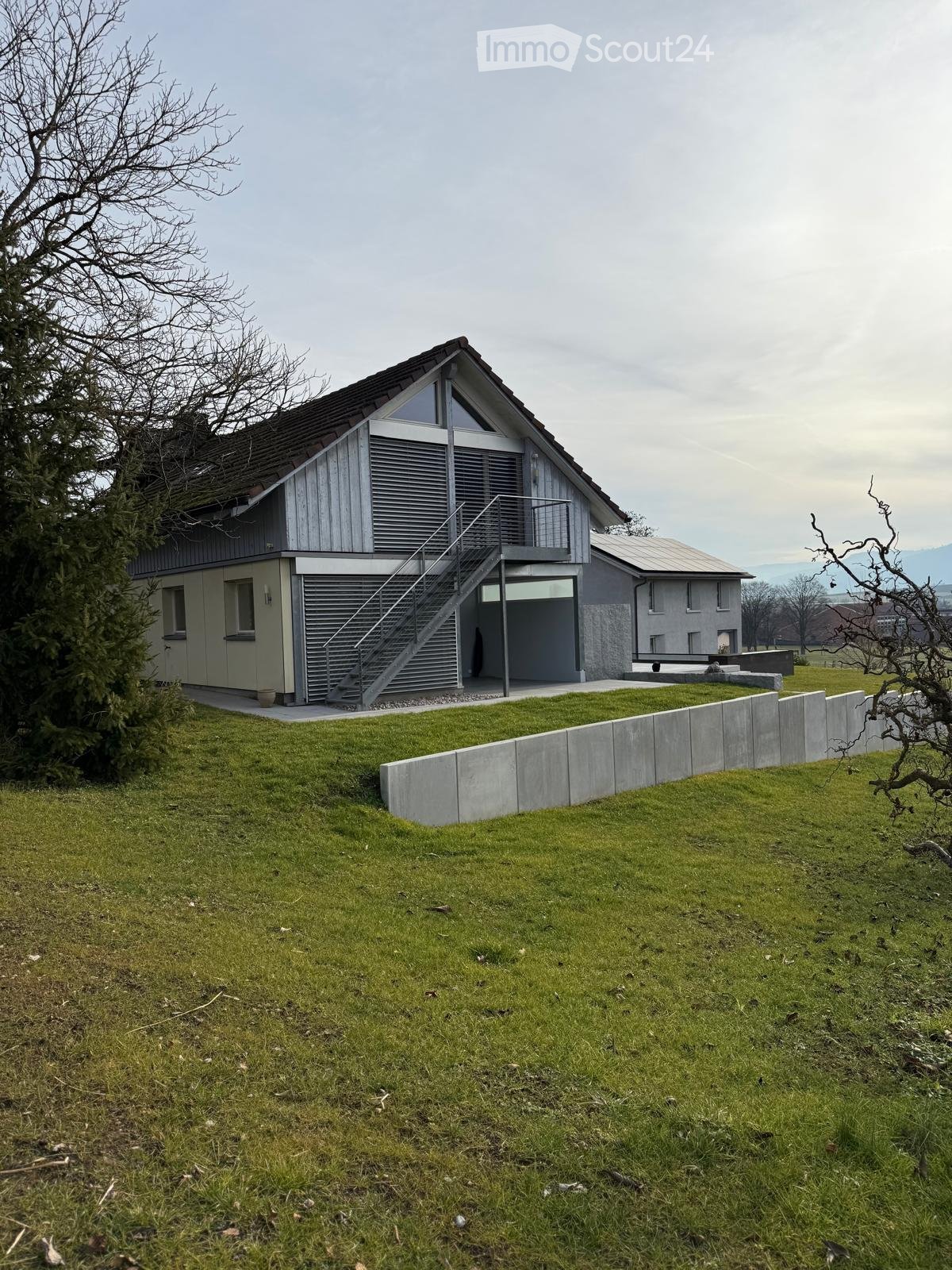 Modern 2 story house, grey and white facade, large windows, gray roof, driveway, large grass field, concrete barrier