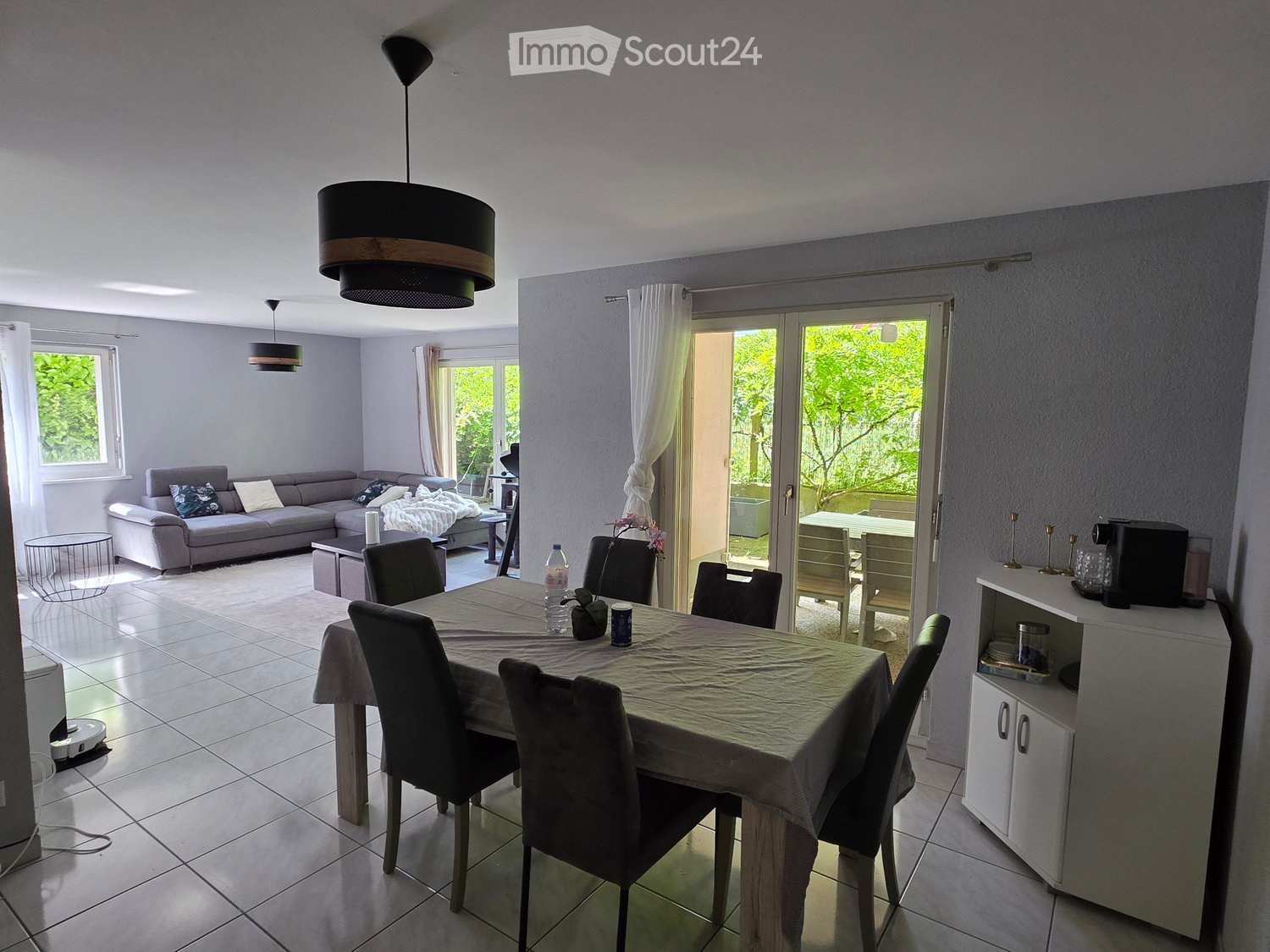 Bright living and dining area with a grey sofa, coffee table, dining table with chairs, and a small cabinet with a coffee machine. Large windows and sliding glass doors lead to a balcony.