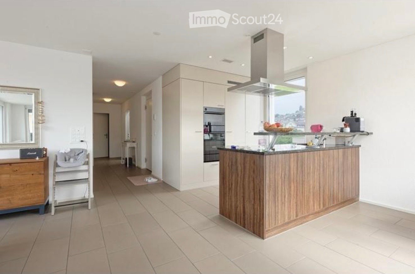 Modern kitchen with a central island, built-in oven, range hood, and a small dining area with a table and chairs. A baby chair and a mirror are also visible.