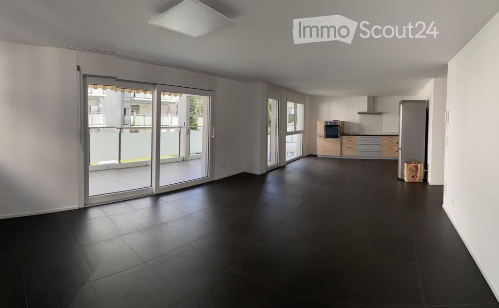 Open-plan unfurnished space with dark tiled flooring, large sliding glass doors leading to a balcony, and a small kitchen area with cabinets and appliances visible in the background.