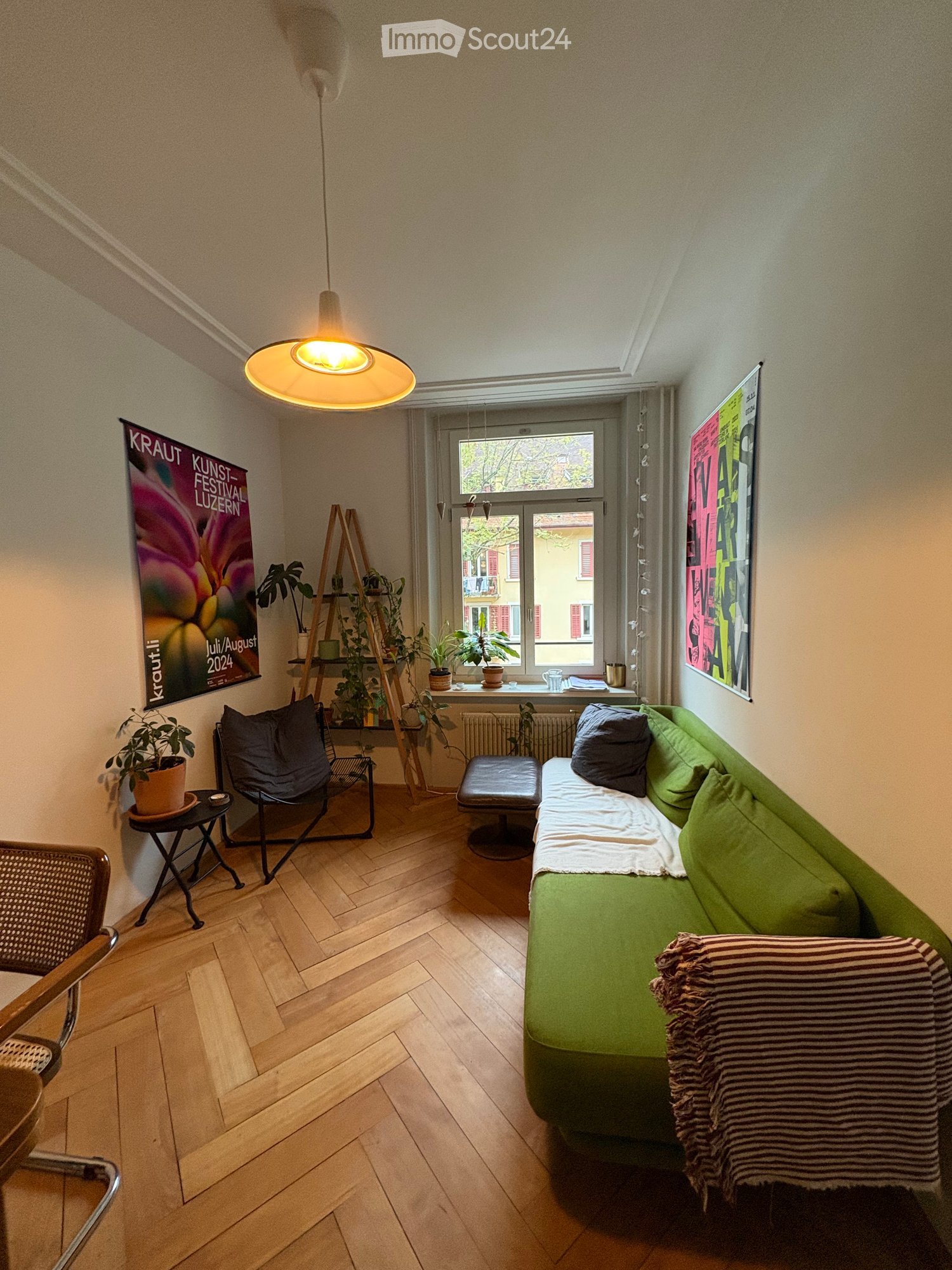 A cozy living room with a green sofa, a black chair, a small table, a stool, potted plants, and posters on the walls. The room features wooden flooring and a hanging lamp.