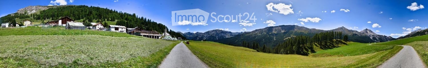 A scenic mountain valley with a winding road, green fields, scattered houses, and distant peaks under a clear blue sky.