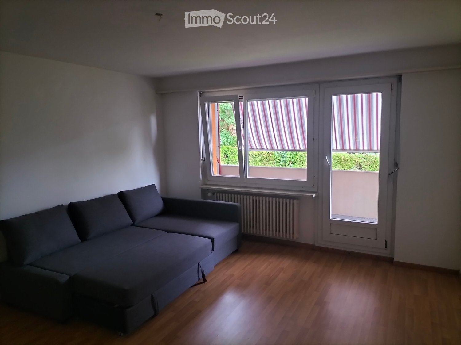 A furnished room with a grey sectional sofa, wooden flooring, and large windows with striped blinds. The room has white walls and a radiator under the windows.