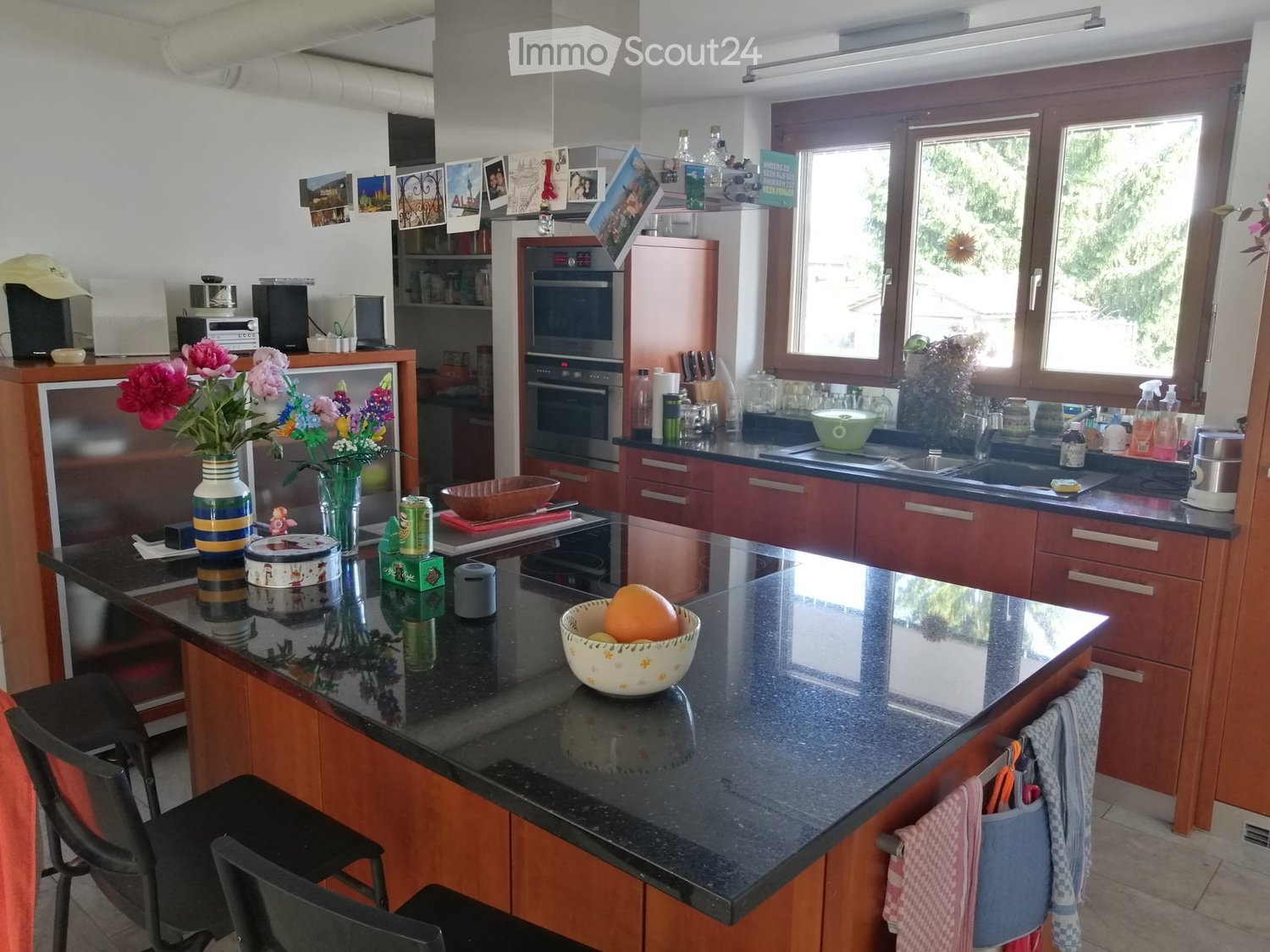 Modern kitchen with wooden cabinets, granite countertop, and various appliances including a refrigerator, oven, and microwave. There is a bowl of fruit on the island, a vase with flowers, and a sink with a faucet.