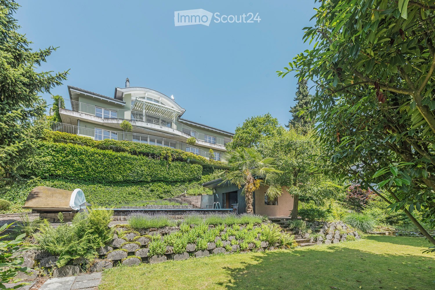 A large, modern house with a curved design and multiple balconies is surrounded by lush greenery, including trees, shrubs, and a well-maintained lawn. A swimming pool is visible in the foreground, along with a stone pathway and a small outdoor seating are