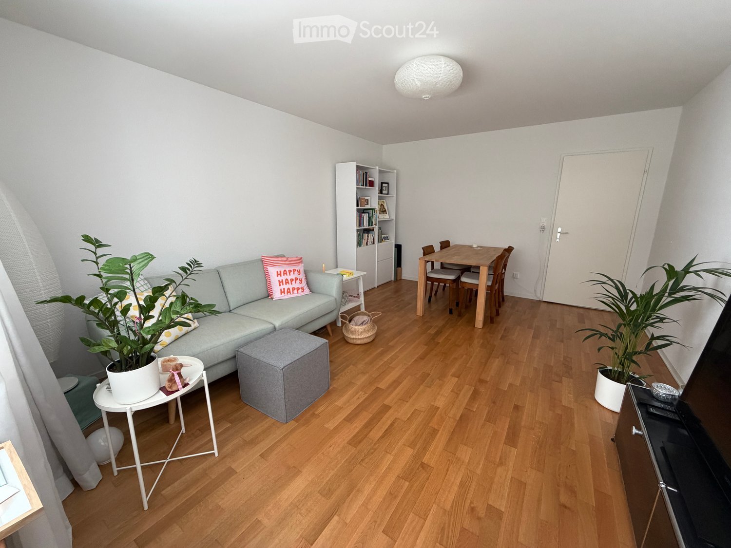A modern living room with a grey sofa, a small round table, a grey ottoman, a dining table with chairs, a bookshelf, and potted plants.