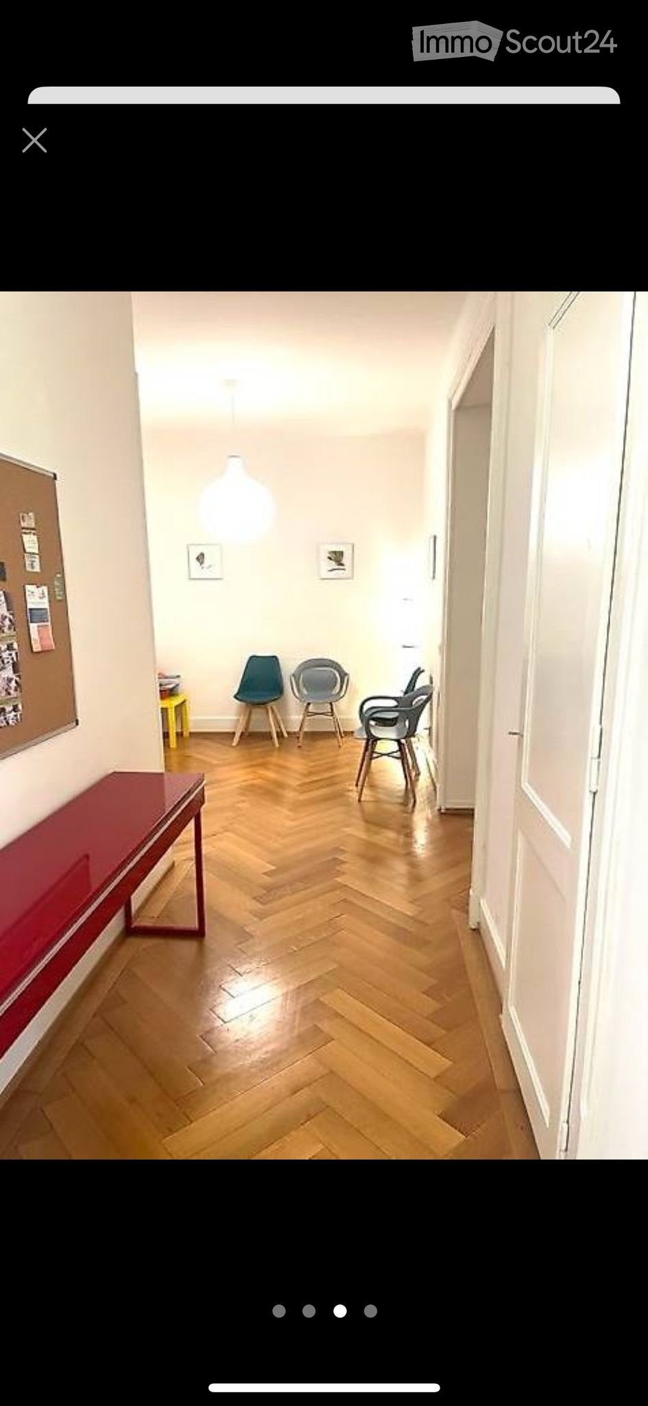 A narrow hallway with wooden herringbone flooring, white walls, and modern lighting. Contains a red bench, a yellow stool, and several chairs.