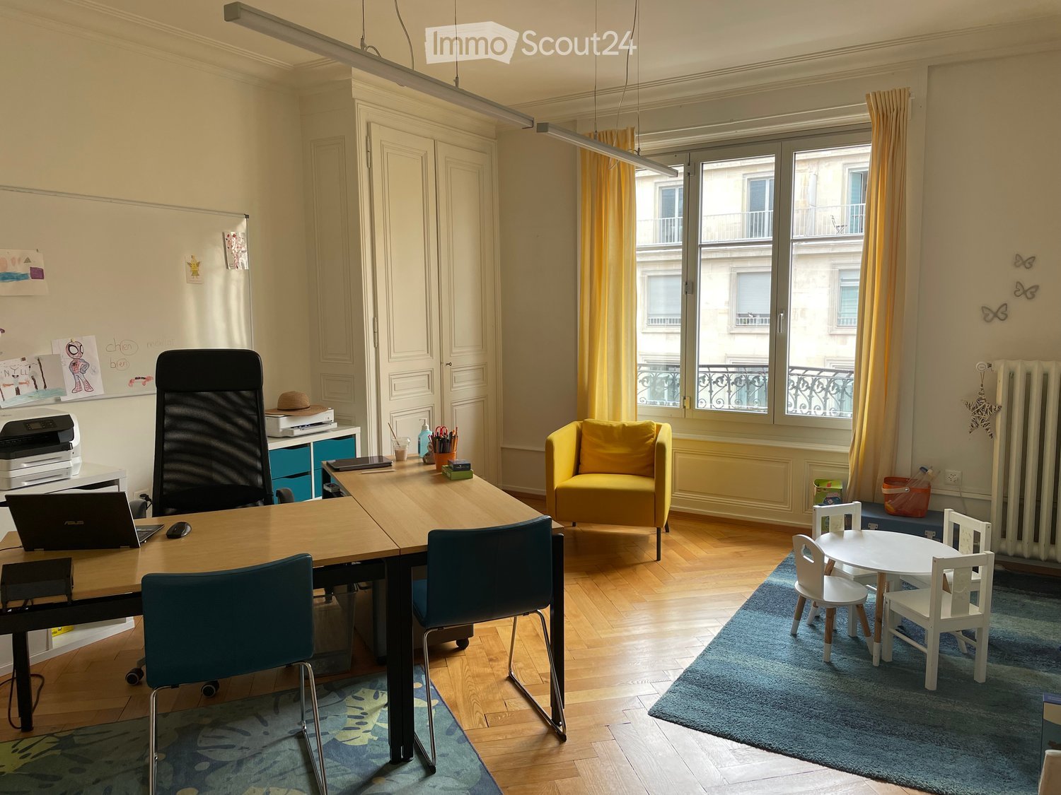 Bright room with wooden flooring, large window with yellow curtains, office area with a table, chairs, laptop, printer, and a yellow armchair. A small play area with a table and chairs is also present.