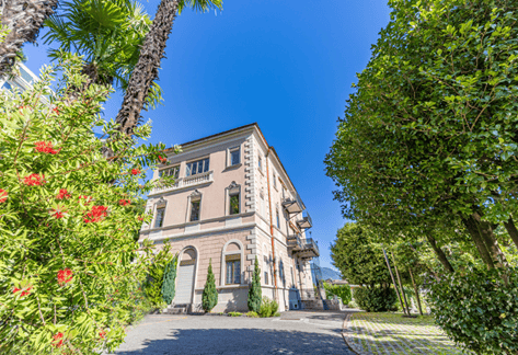 A large, elegant villa with classical architecture, surrounded by lush greenery and palm trees. The building features arched windows, a symmetrical facade, and a well-maintained garden.