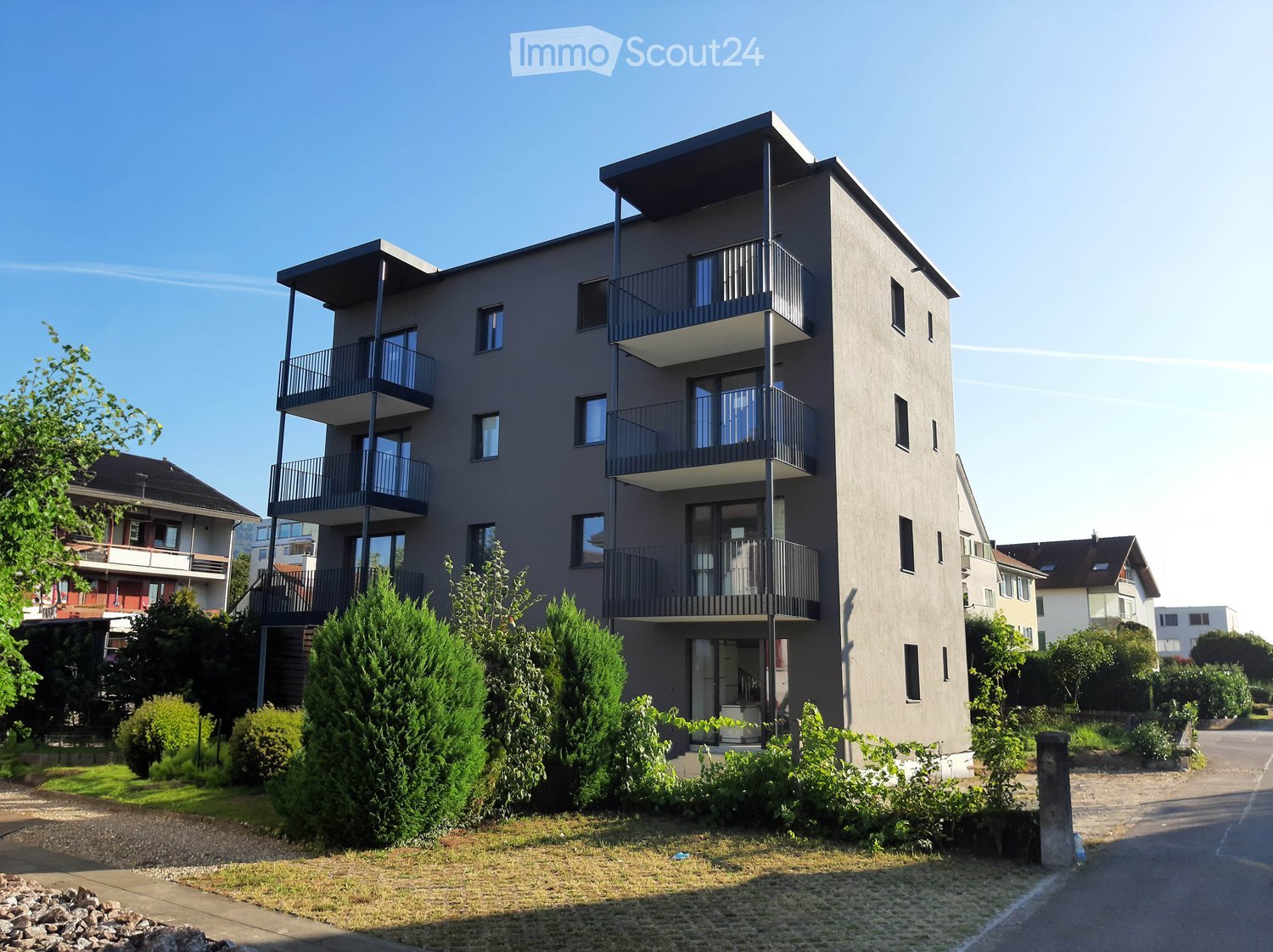 multi-story apartment building, gray exterior, balconies, small garden in front, clear blue sky