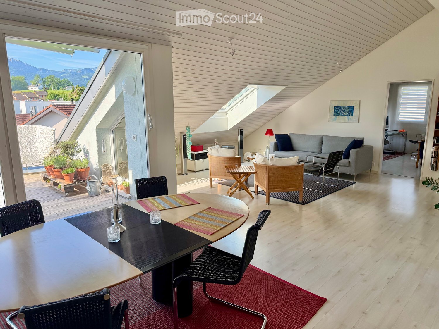 Bright living room with wooden floors, a grey sofa, a round dining table with chairs, and a view of the mountains through large windows.