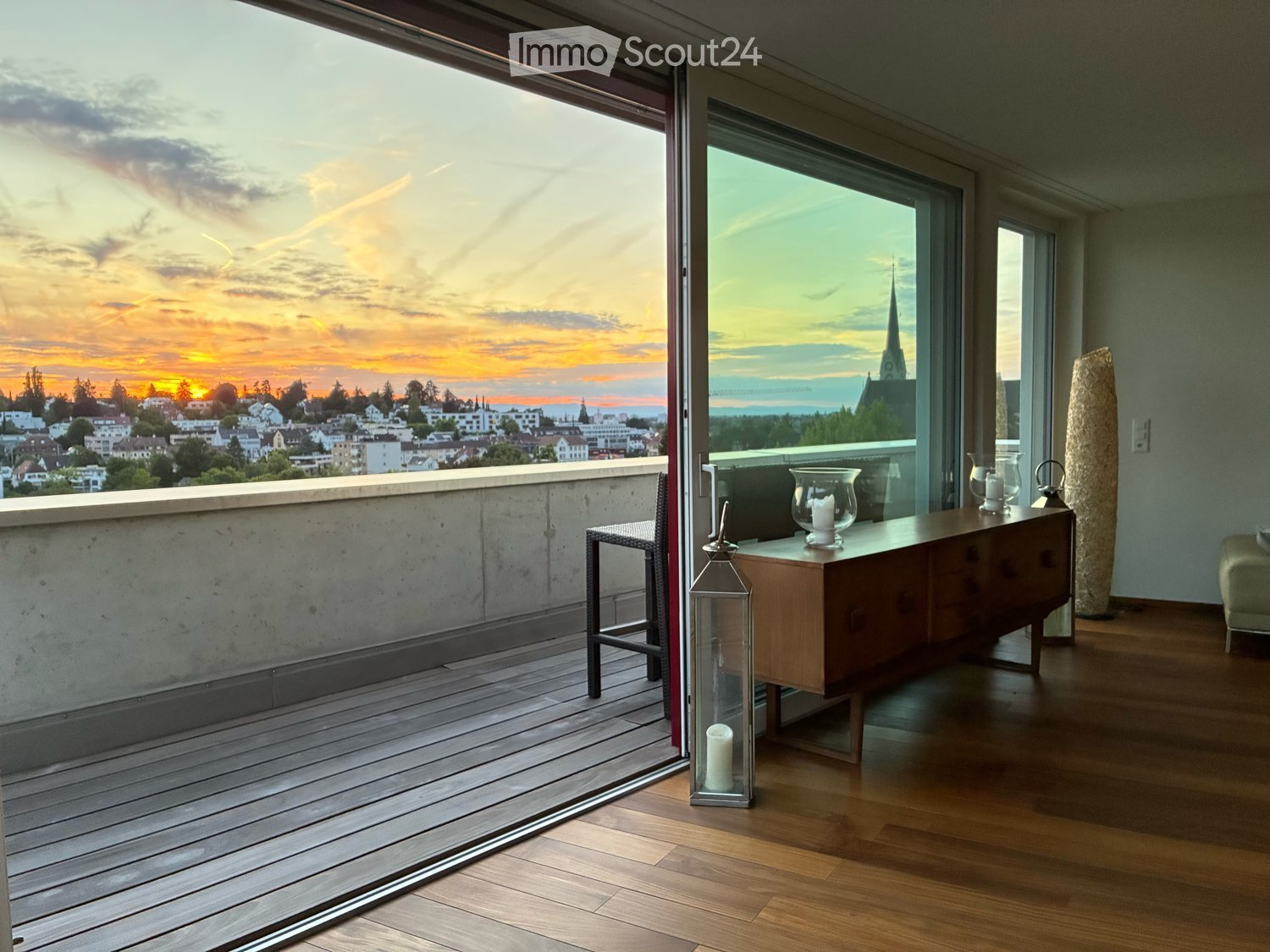 Balcony with wooden decking, modern furniture, and a scenic city view at sunset.
