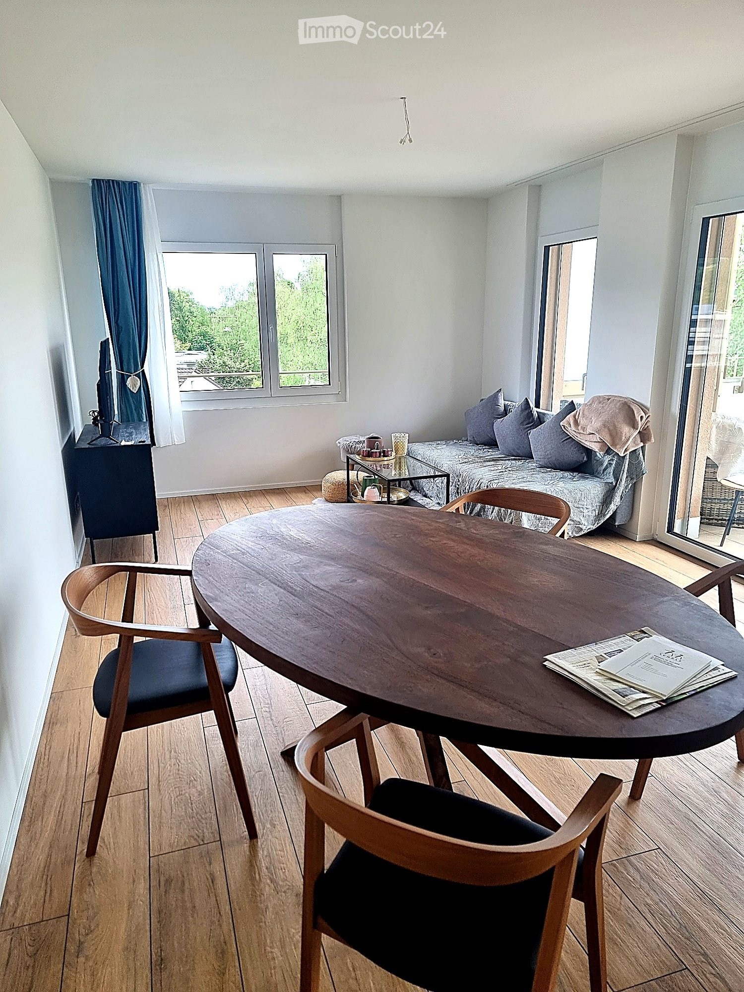 A modern, well-lit room with wooden flooring, a round wooden table with chairs, a sofa with pillows, a television on a stand, and a small side table with cups and a stool.
