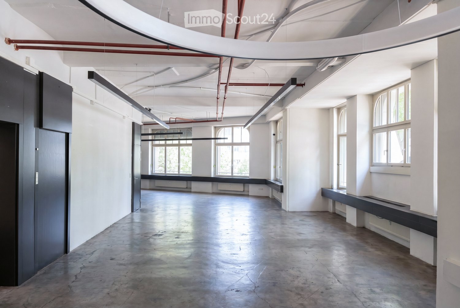 An empty, industrial-style room with white walls, concrete floor, large windows, and exposed pipes and lighting fixtures.