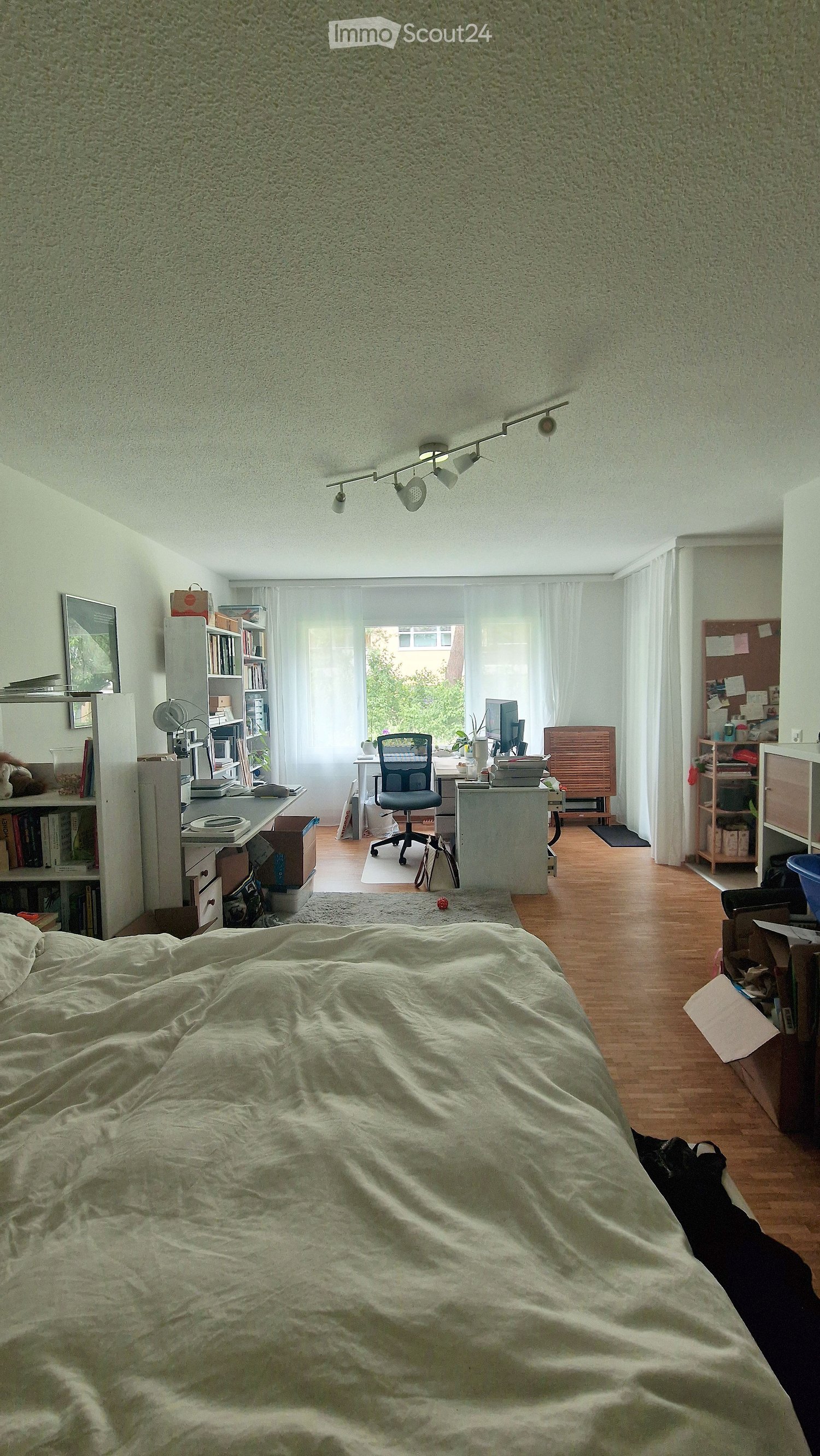 Cozy bedroom with a large bed, wooden floor, large window with white curtains, desk with chair, bookshelf, and a lamp.