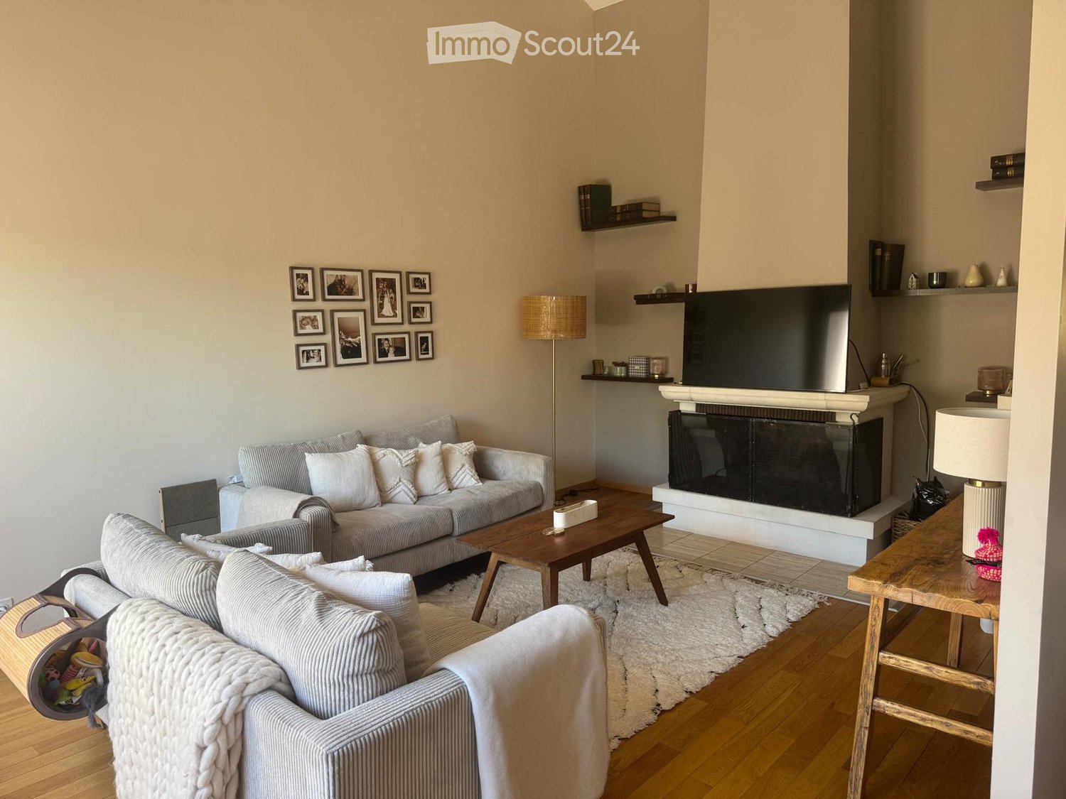 Modern living room with a grey sofa, armchair, coffee table, fireplace, floor lamp, and shelves with picture frames.