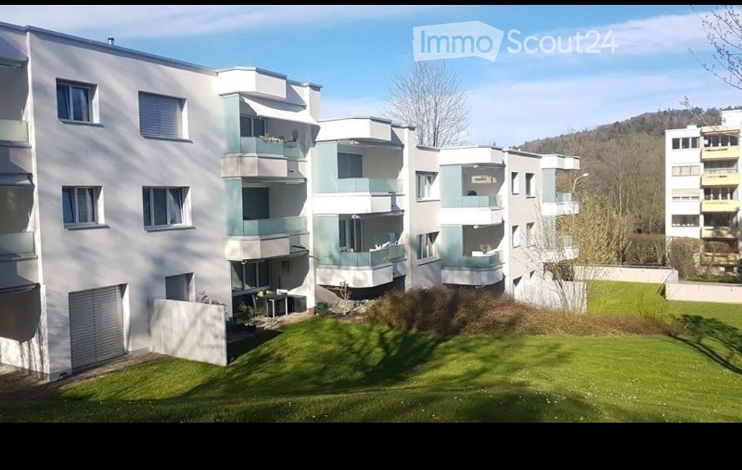 Modern multi-story residential building with white and light green facade, multiple balconies, and a well-maintained lawn in front.