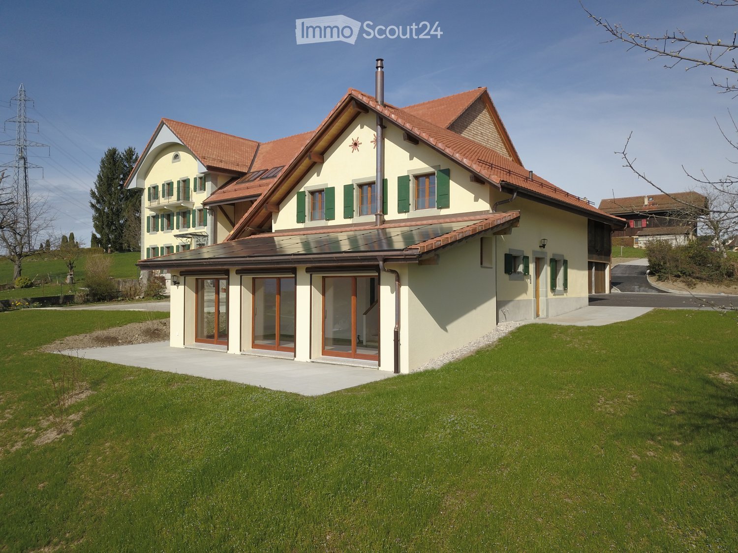 Two-story house with a red-tiled roof, green shutters, and a modern extension with large glass doors. The house is surrounded by a well-maintained lawn and is situated in a rural area with other houses and trees in the background.