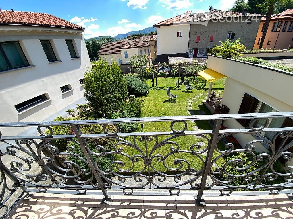Balcony overlooking a well-maintained garden with lawn, trees, and outdoor furniture, surrounded by residential buildings and a scenic mountain view.