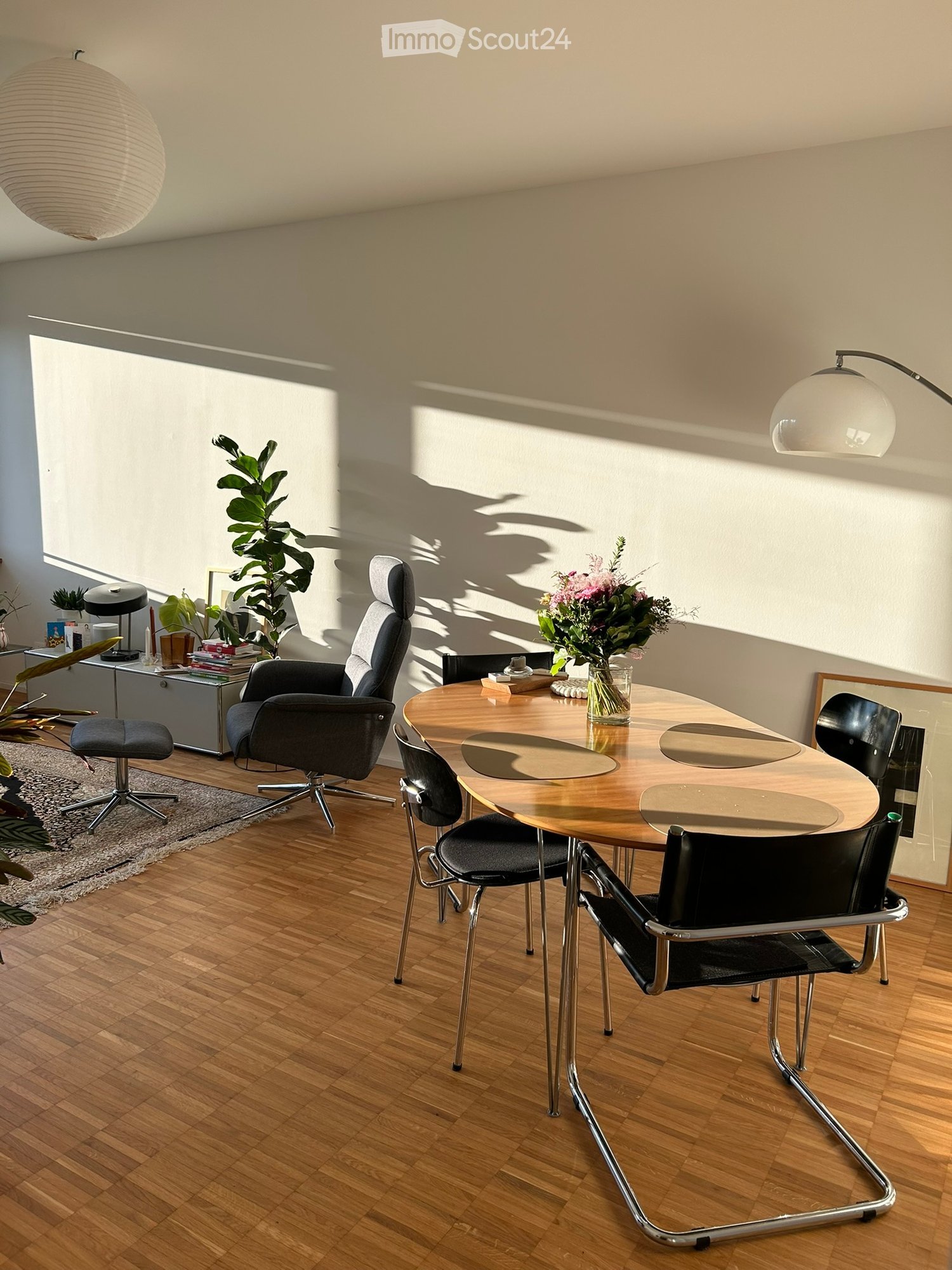 Modern dining area with a round wooden table, black chairs, a grey leather chair, a floor lamp, a plant, and decorative items on a side table.