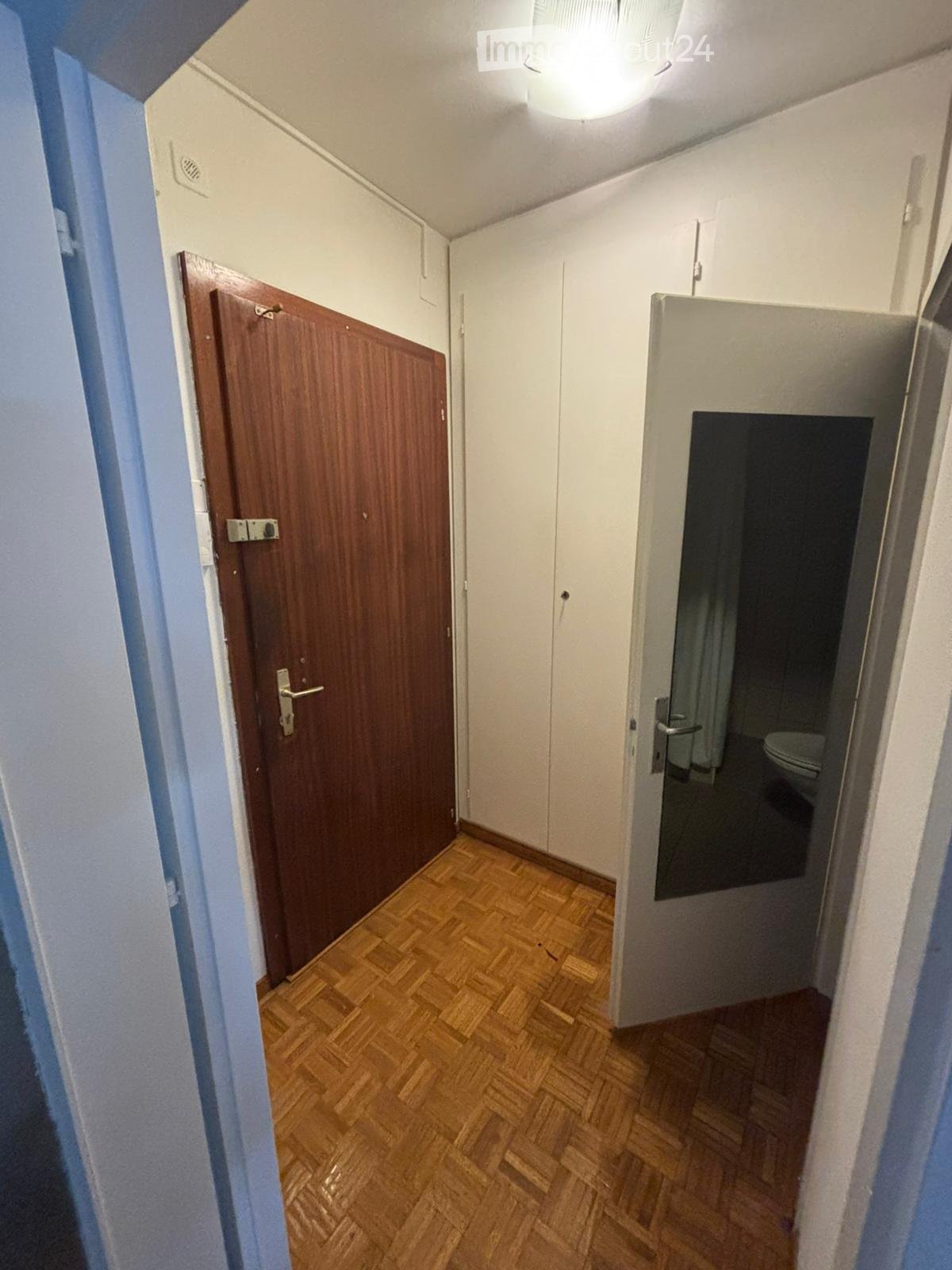 A narrow hallway with wooden flooring, white walls, and a wooden door on the left leading to another room. A partially open door on the right reveals a bathroom with a toilet and a mirror.