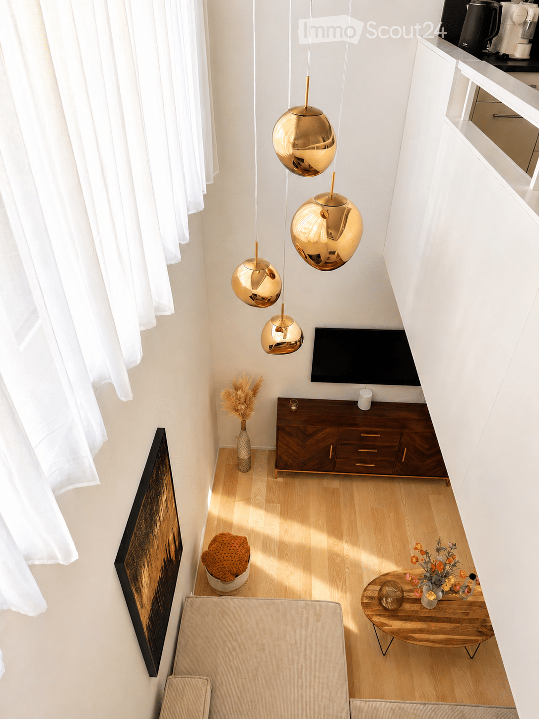Modern living room with light wooden flooring, white walls, and large windows with white curtains. Features a beige sofa, wooden coffee table with a vase of flowers, TV stand with a flat-screen TV, and decorative items including a woven basket and a vase