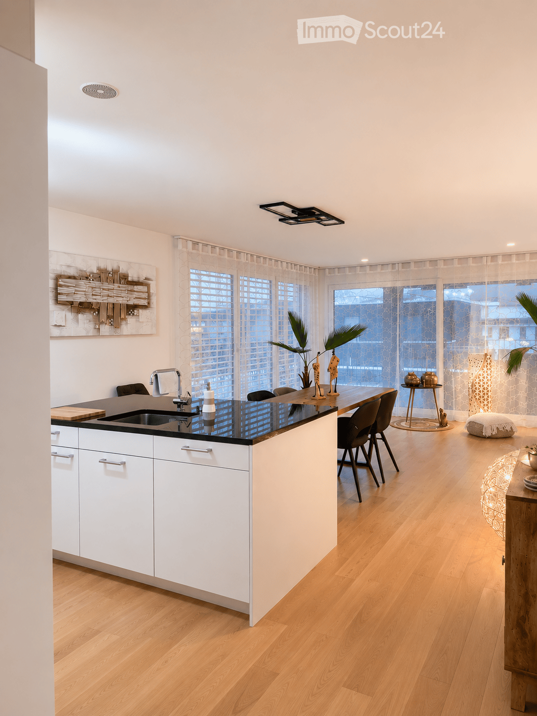 Modern kitchen with white cabinets, black countertop, island, sink, and dining area with chairs and table. Features large windows with sheer curtains, wooden flooring, and decorative items including a plant and lamp.