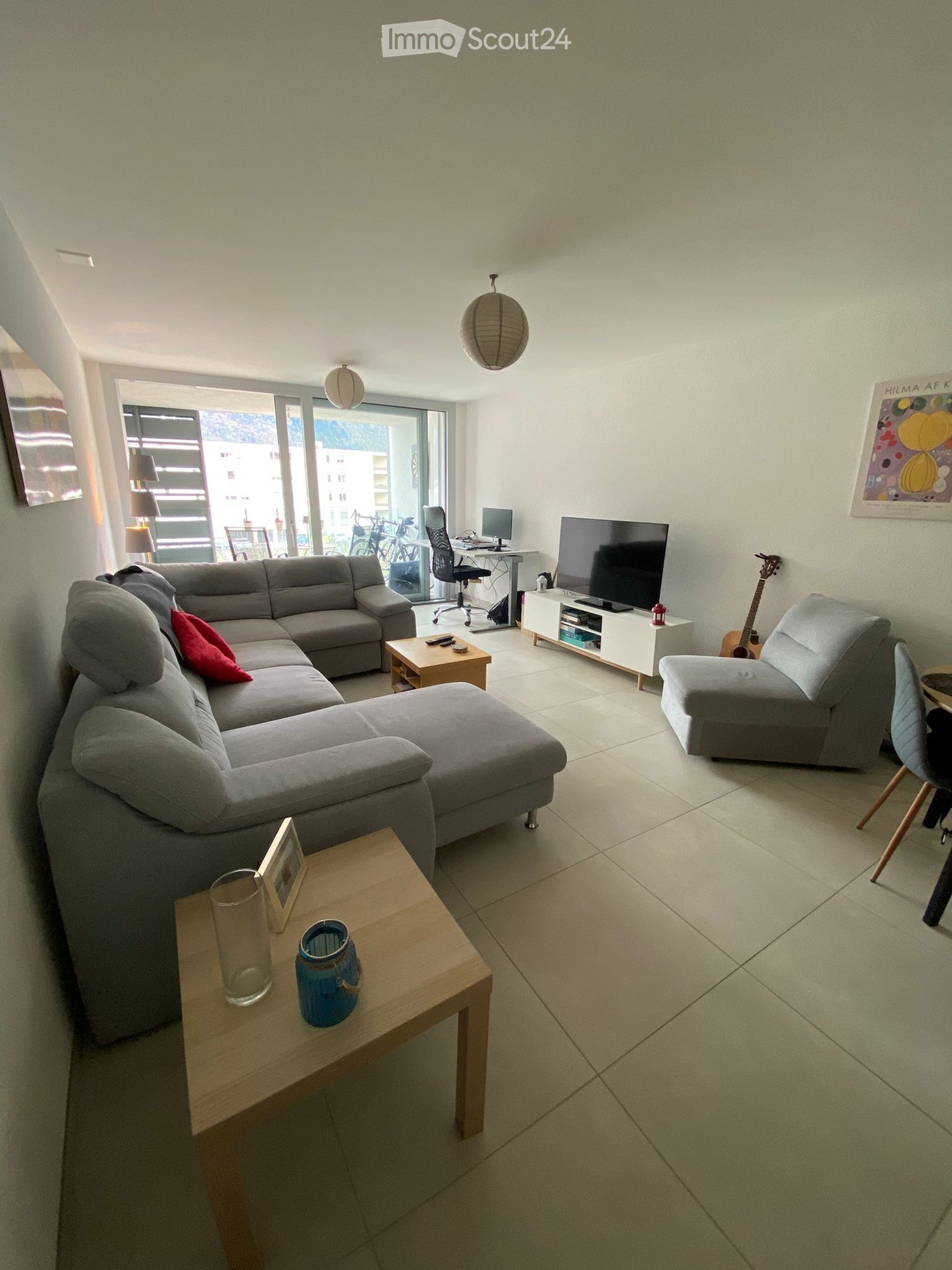 Modern living room with a grey sectional sofa, coffee tables, a guitar, a TV, and a view of the city through large windows.