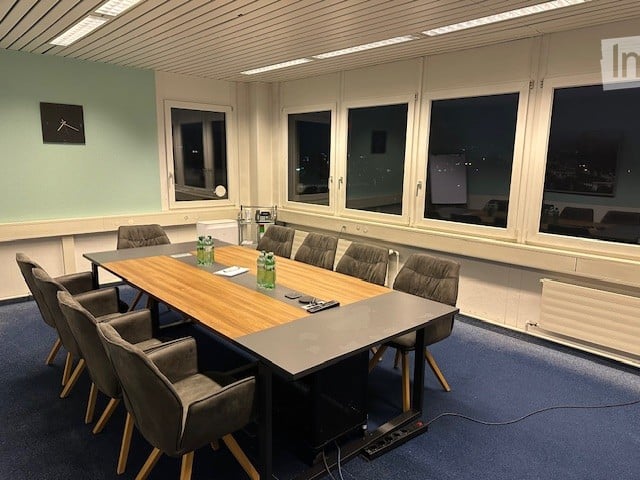 Long dining table, 8 chairs around, projector, clock on wall, blue floor, white walls, large windows