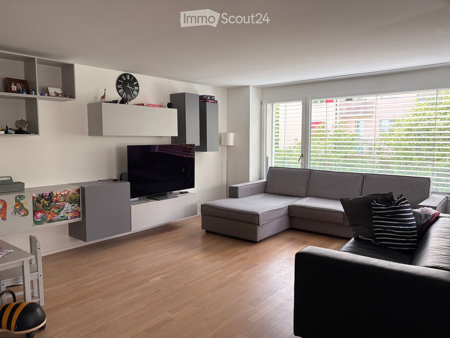 Modern living room with a grey sectional sofa, coffee table, flat-screen TV, wall-mounted shelves, and a floor lamp. The room features wooden flooring and large windows with blinds.
