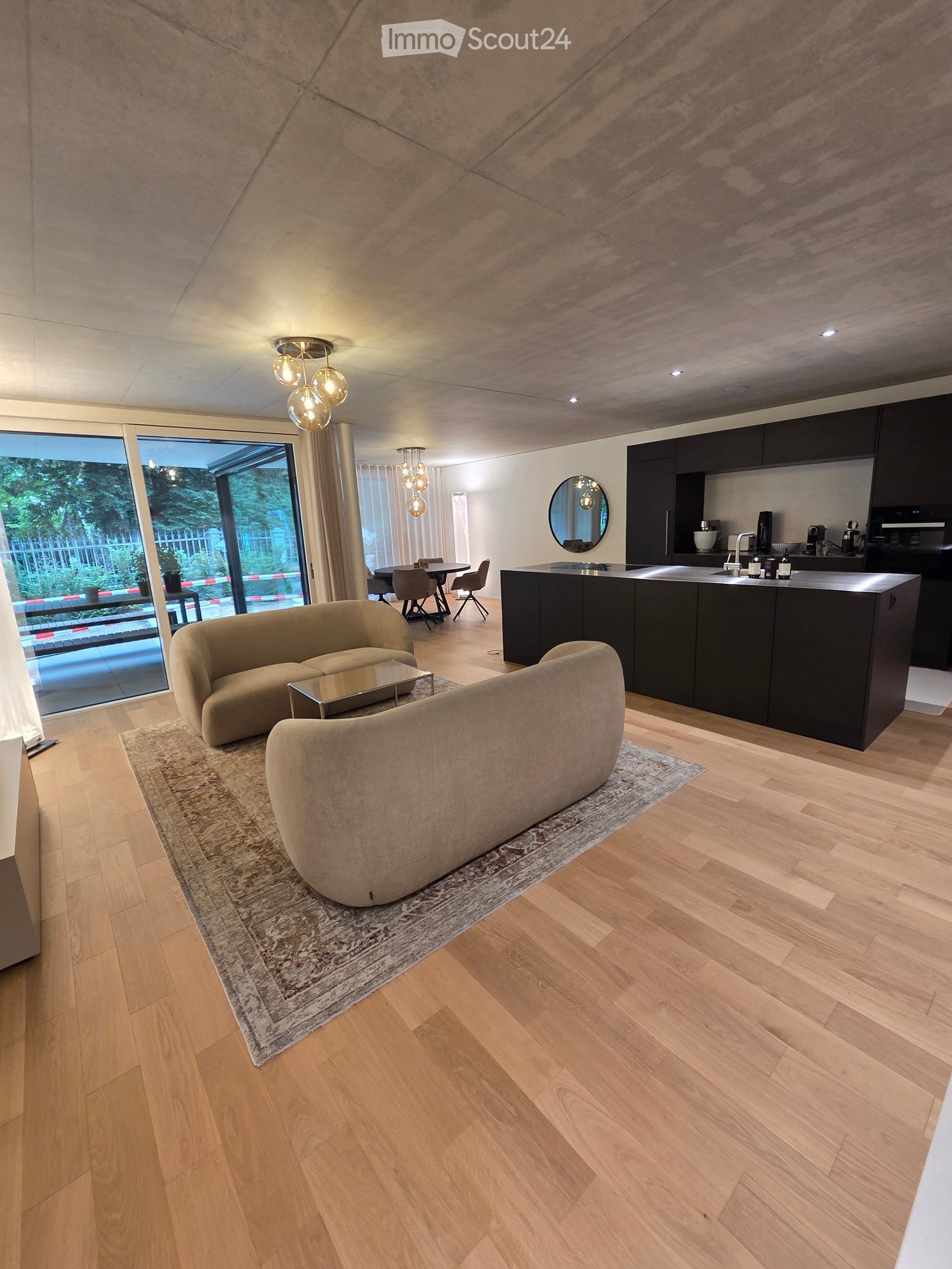 Modern living and dining area with a kitchen island, beige sofa, coffee table, dining table, and chairs. The space features wooden flooring, large windows, and contemporary lighting.