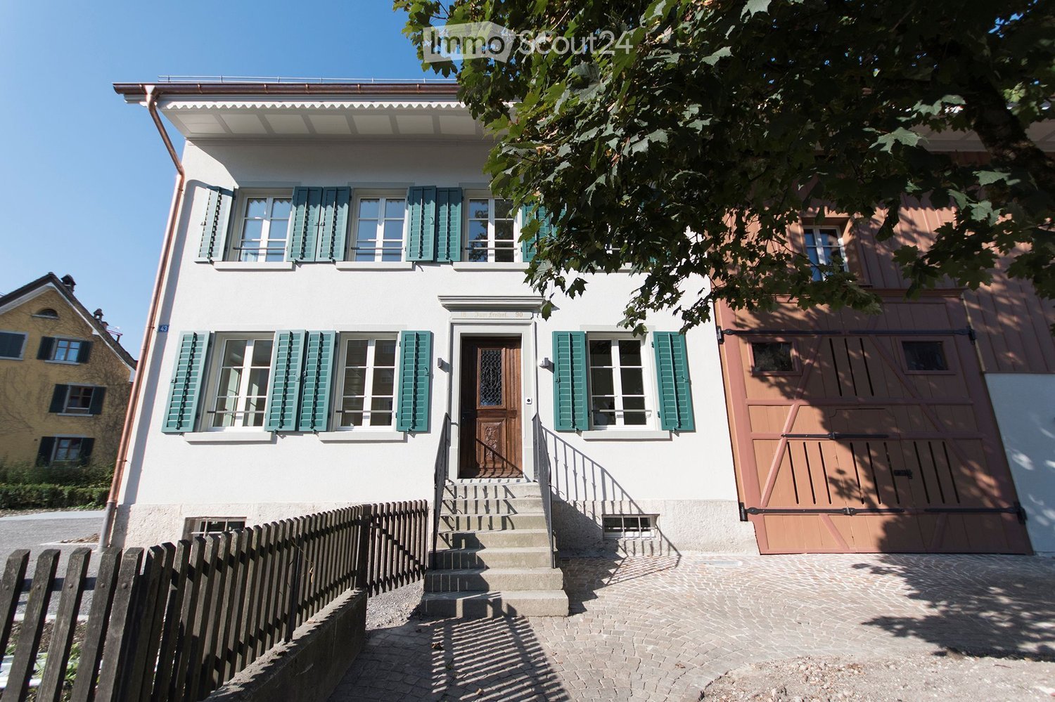 Two-story house with white facade, green shutters, wooden garage door, and steps leading to the entrance.