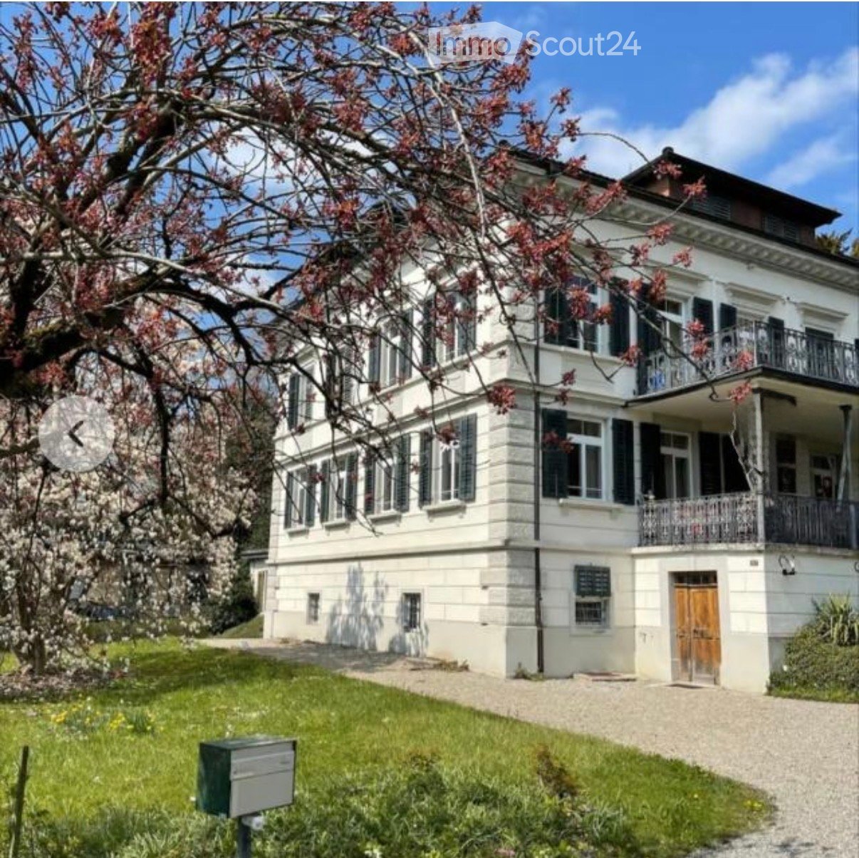 A two-story white house with multiple windows, a balcony, and a wooden front door, surrounded by a well-maintained lawn and blooming trees.