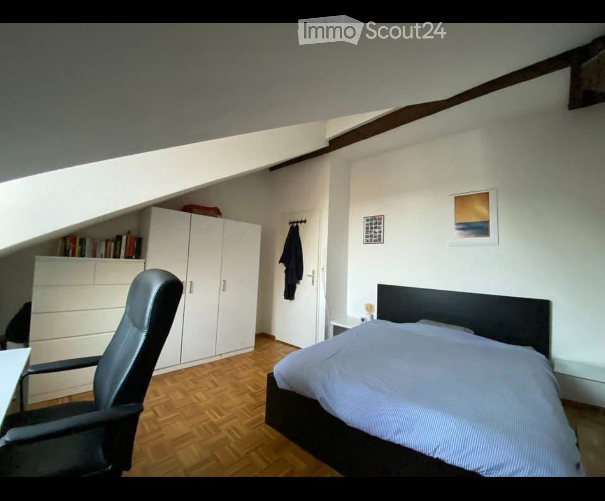 Cozy bedroom with wooden flooring, a bed with a dark headboard, a black office chair, a white wardrobe, and a bookshelf with books.