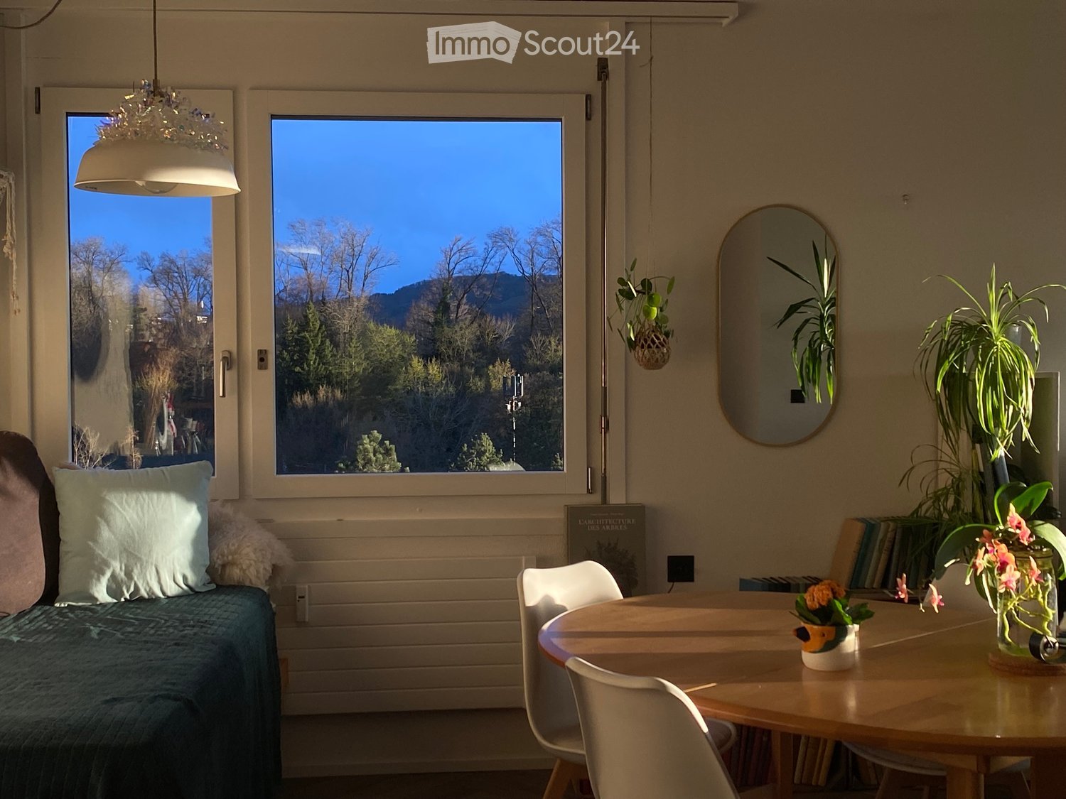 Bright living space with a sofa, round wooden table, chairs, and decorative plants, overlooking a scenic mountain view.