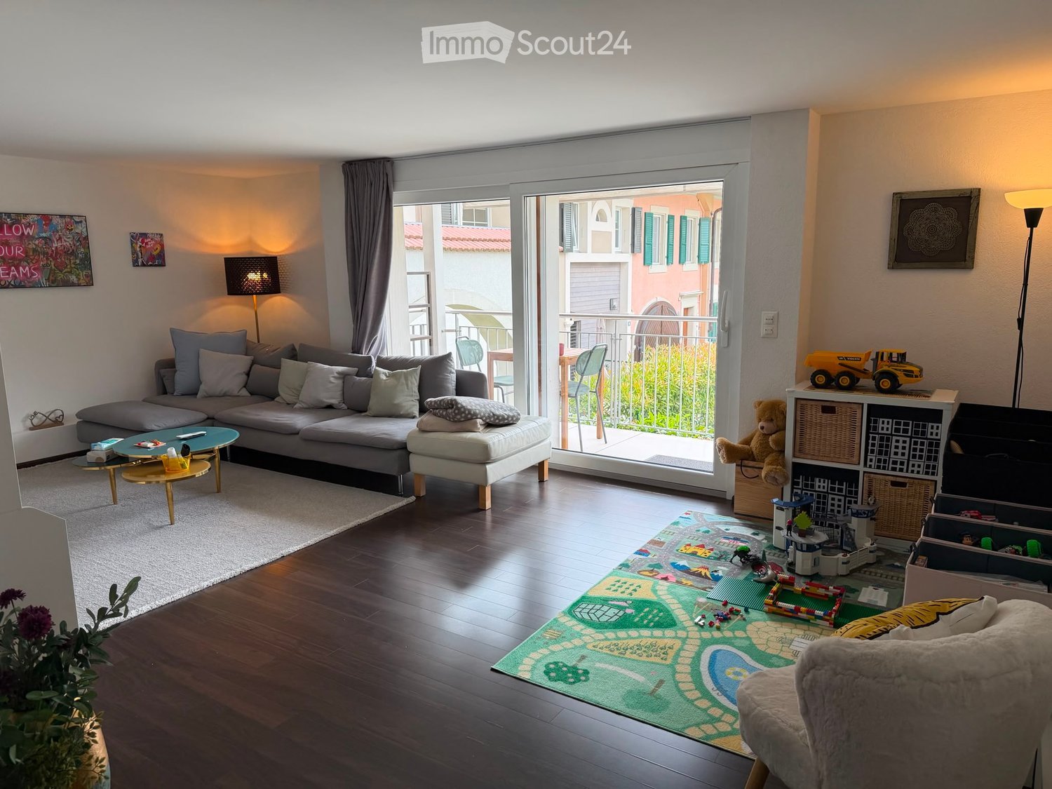 Bright living room with a grey sectional sofa, coffee table, ottoman, floor lamp, and a balcony with outdoor seating. A play area with toys and a storage unit is visible.