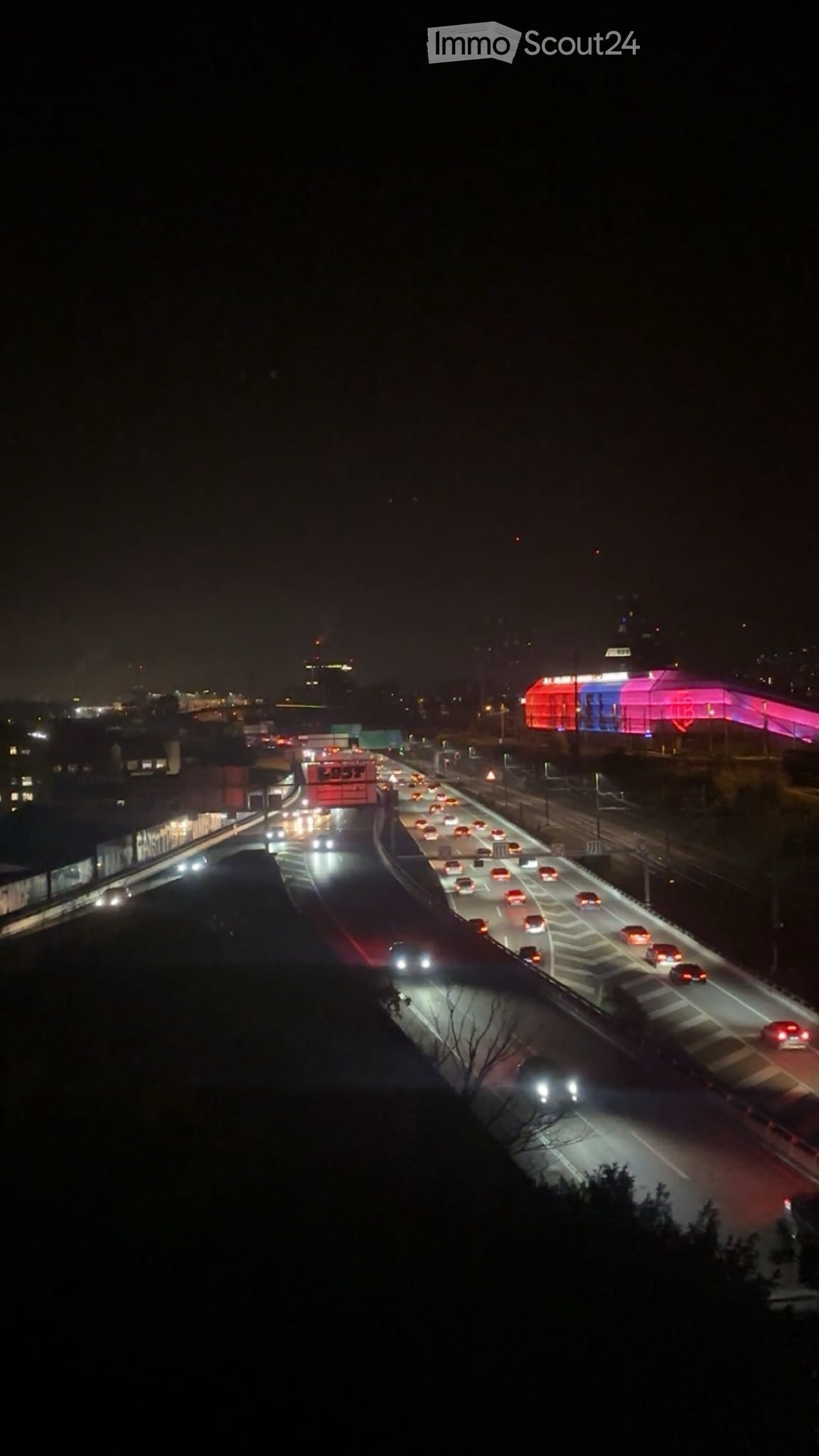 Nighttime aerial view of a busy highway with multiple lanes of traffic, illuminated vehicles, and distant city lights.
