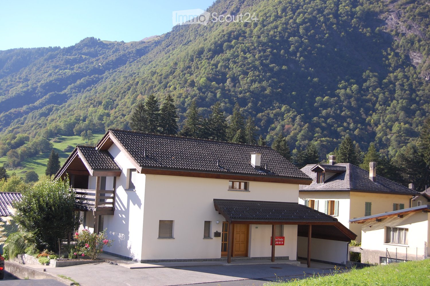 A two-story house with a dark tiled roof, white walls, and a wooden balcony on the left side. The house has multiple windows and a covered entrance with a wooden door. The surrounding area includes a driveway, a small garden with flowers, and a scenic mou
