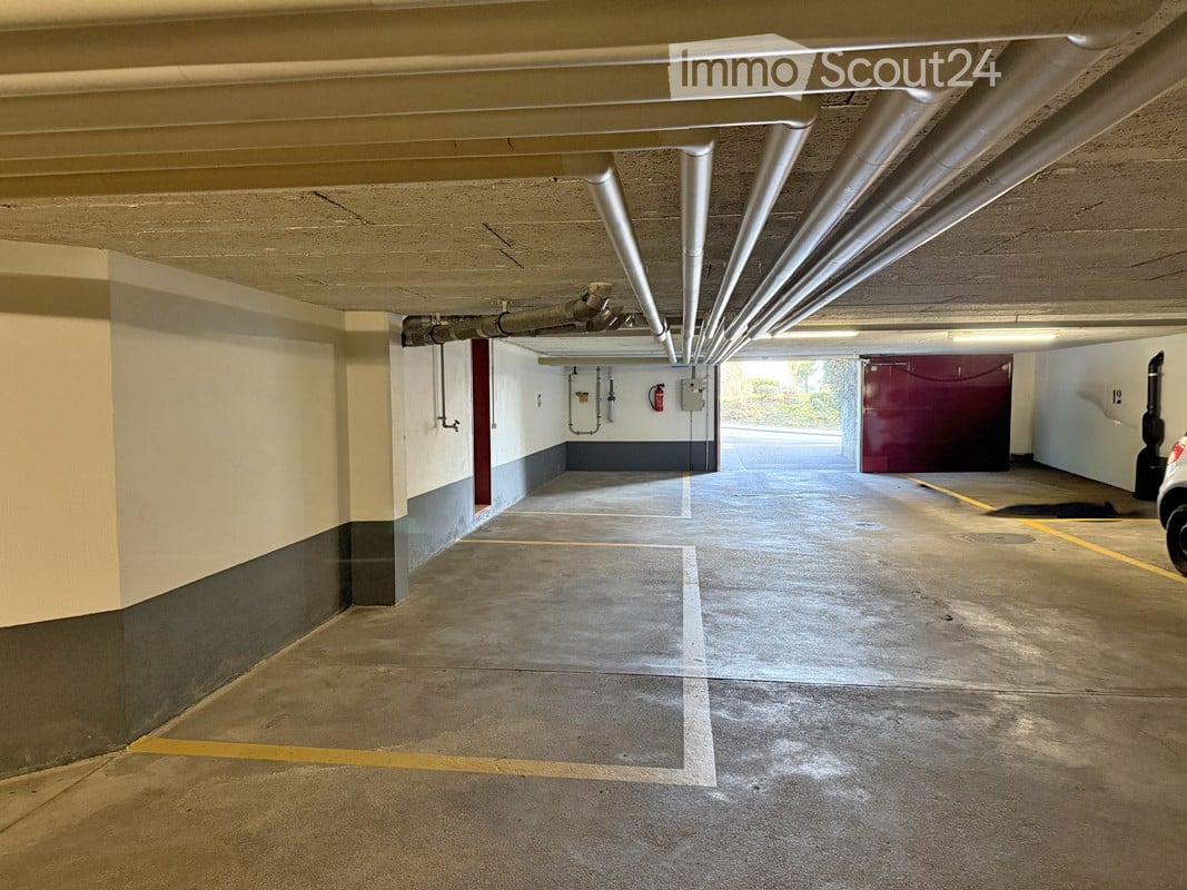 Underground parking garage with marked parking spaces, concrete floors, and exposed pipes. A red storage unit is visible in the background.