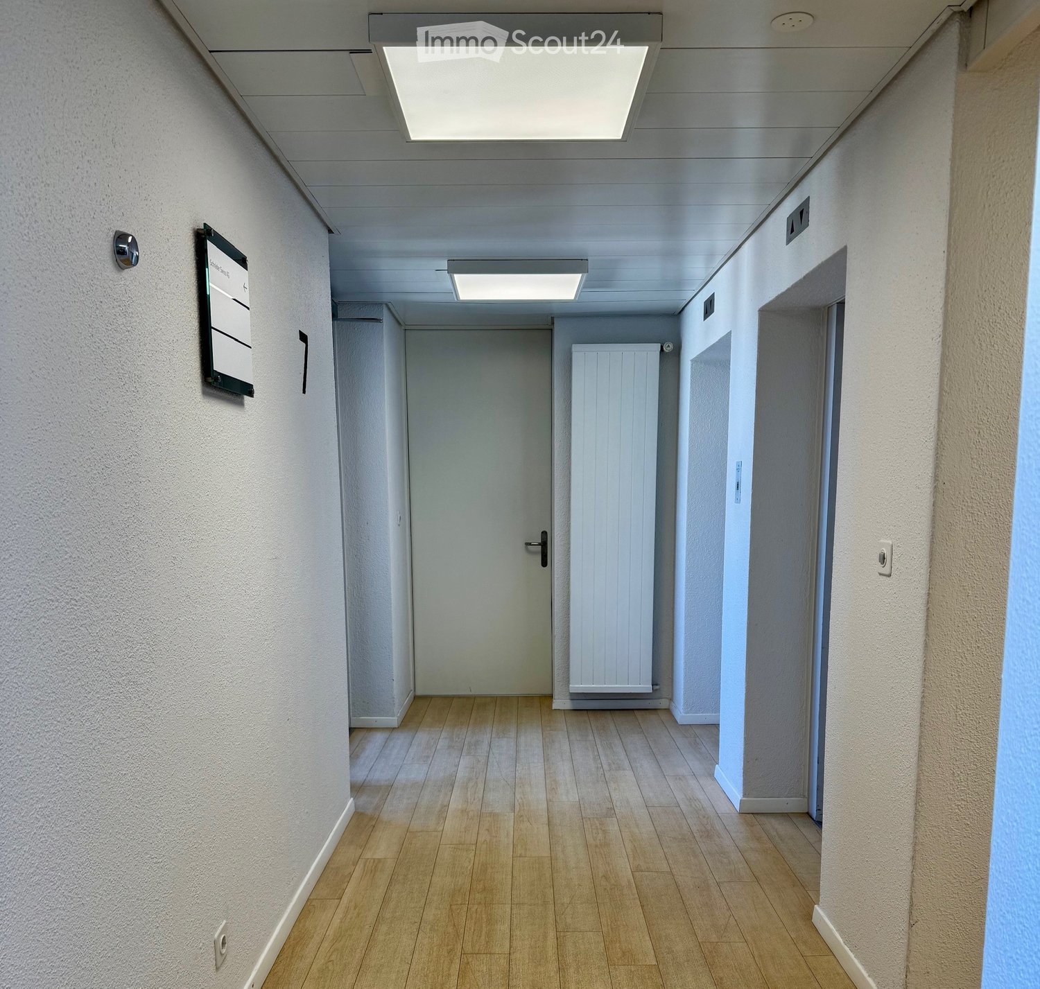 Clean, well-lit hallway with wooden flooring, white walls, and a closed door at the end. Features a radiator on the right wall and a signboard on the left wall.