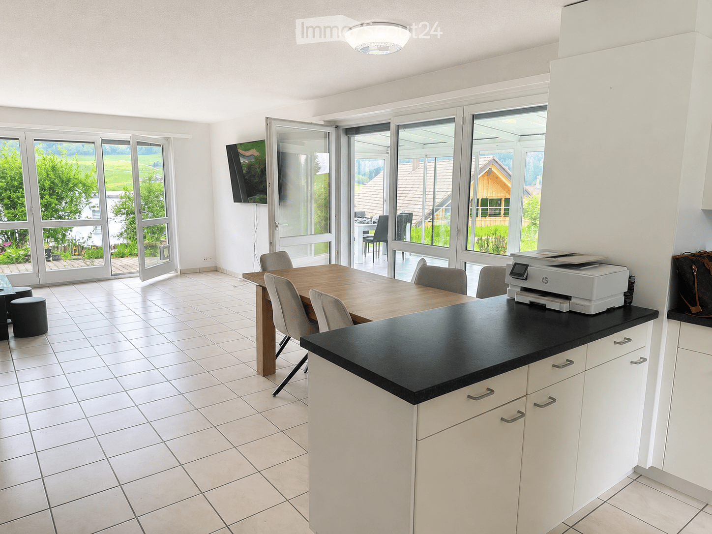 Bright kitchen with white cabinets, black countertop, tiled floor, and large windows overlooking a green landscape. Includes a dining table with chairs, a printer on the counter, and a TV mounted on the wall.