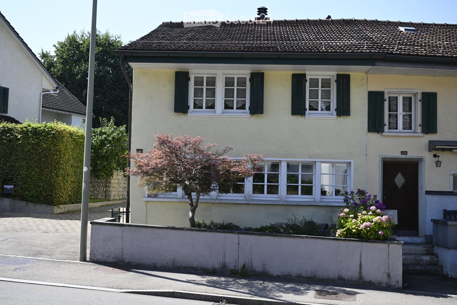 Two-story house with light yellow walls, dark green shutters, multiple windows, and a small garden with flowers and a tree.