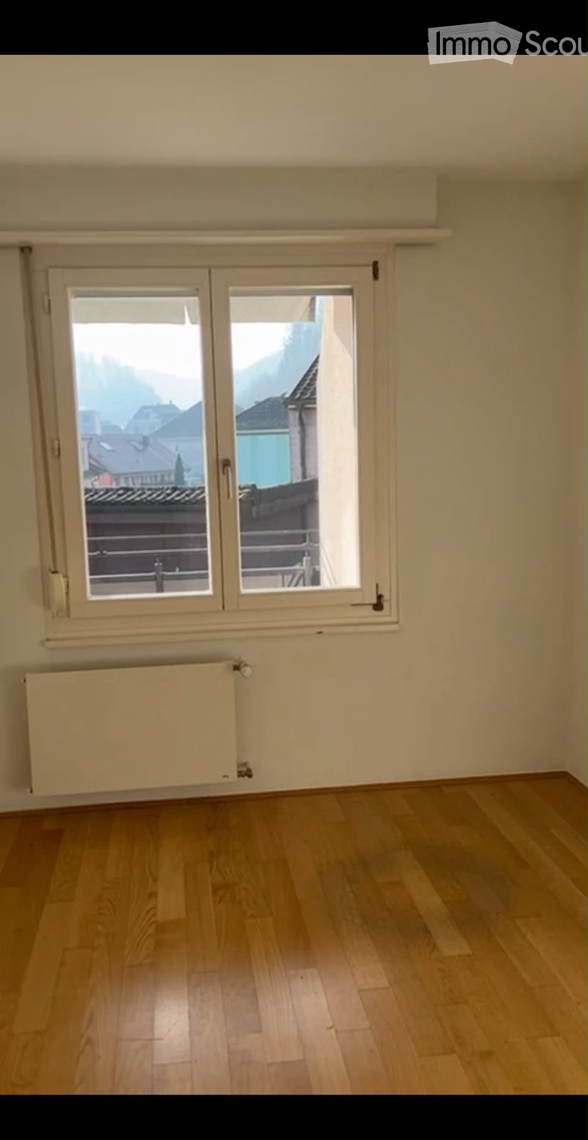 Empty room with wooden flooring, large window with double doors leading to a balcony, radiator below the window, and a view of rooftops and mountains.