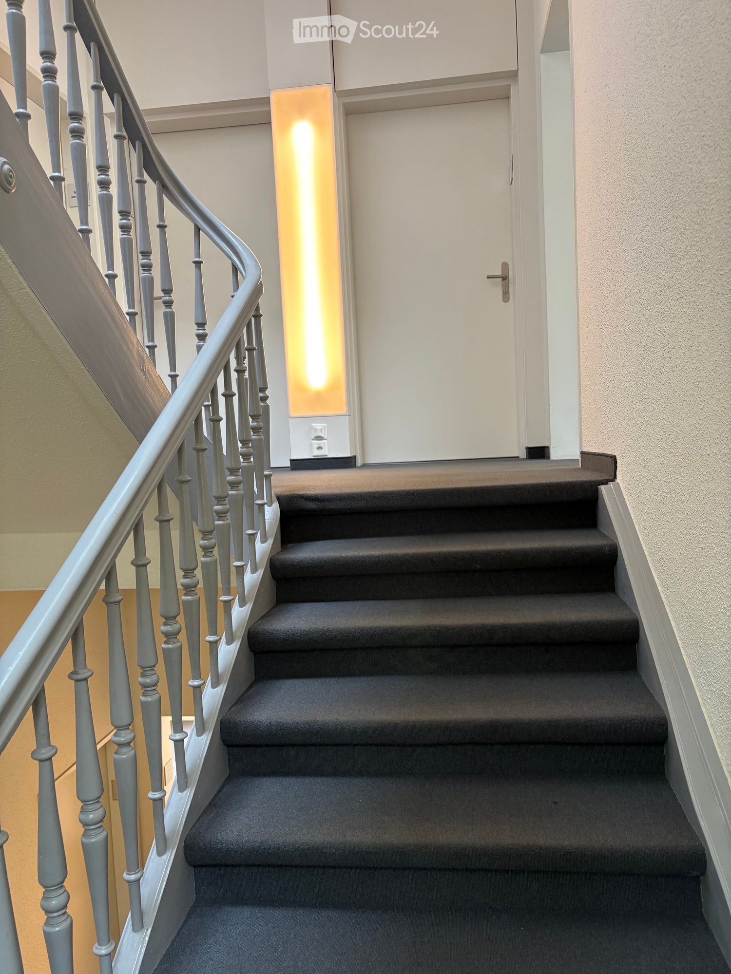 Spiral staircase with dark carpeted steps and a grey metal balustrade, leading to a closed white door with a lit panel beside it.