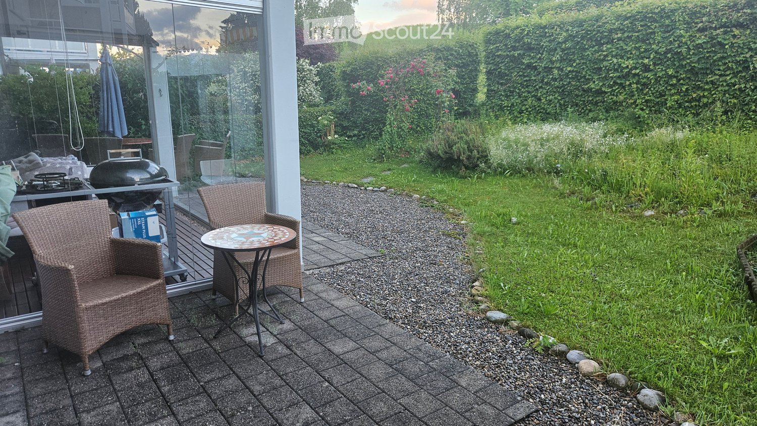 Outdoor patio area with wicker chairs, a small table, a grill, and an umbrella near a glass-enclosed space. The area is bordered by a gravel path and a well-maintained lawn with hedges and flowering plants.